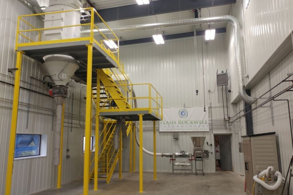 Interior of a Flash Rockwell facility showing a compact industrial flash dryer with small footprint mounted above a bright yellow platform and stairs, connected to ducts and piping, with a “Flash Rockwell Technologies” banner on the back wall