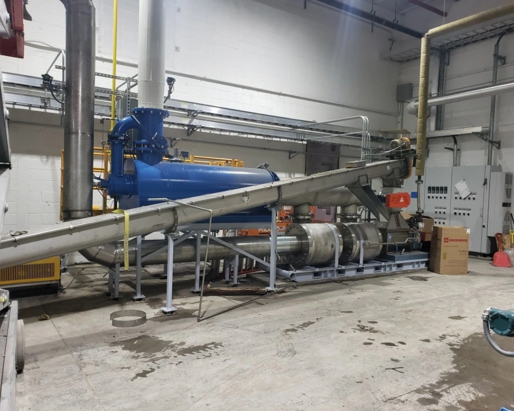 Flash Rockwell low pressure flash drying system installed indoors, showing a large horizontal blue dryer connected to an inclined stainless-steel conveyor and piping, with control panels, support frames, and plant equipment in the background.